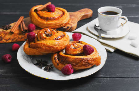 Round twisted Denish Buns with berries raspberries, syrup, white cup of espresso coffee, sugar cubes, cinnamon sticks, star anise, cutting board made of wood olive on a black wooden backgroundの写真素材