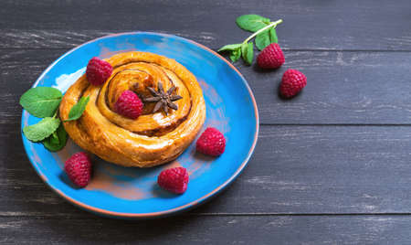Denish Bun with raspberry, cinnamon, star anise, mint on a blue ceramic plate worn over dark black wood background, empty place for your textの写真素材