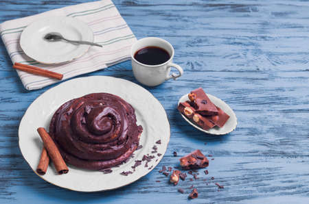 Round Twisted Bun Danish snail poured chocolate icing, white cup of coffee espresso, cinnamon sticks, pieces of chocolate with hazelnuts on a blue wooden background, empty clean place for your textの写真素材