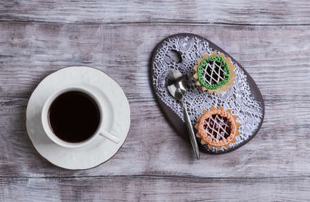 Small cakes petit fours tartlets with jam and cream, ceramic board with a pattern of white lace, with a cup of coffee espresso on a light wooden background, top viewの写真素材