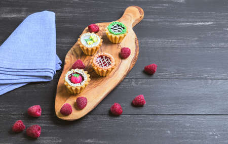 Small cakes petit fours in the form of baskets with cream and jam on a wooden plank from a tree of olive, blue cloth, worn on a dark black background in rustic style, Empty place for textの写真素材