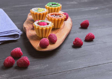 Small cakes petit fours in the form of baskets with cream and jam on a wooden plank from a tree of olive, blue cloth, worn on a dark black background in rustic style, Empty place for textの写真素材