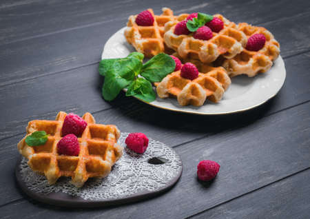 Belgian lush round waffles with fresh raspberries, twigs and leaves mty in a white plate and ceramic board with lace pattern on dark wooden background, empty space for your textの写真素材