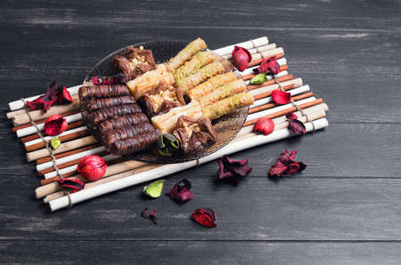 The glass plate and an assortment of baklava and chocolate with nuts, dried flowers red, mat of bamboo on a dark black background in rustic styleの写真素材