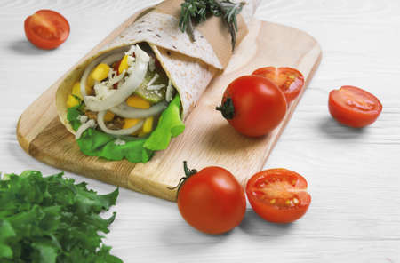 Burrito wrapped in tortilla bread with cereals, corn, onion rings, lettuce, cheese, rosemary, pickled cucumbers, ketchup on a chopping board on a light white wooden backgroundの写真素材