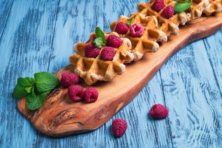 Thick lush Belgian waffles, fresh raspberries and sprigs of mint on a cutting board made of wood olive on a blue wooden background surfaceの写真素材