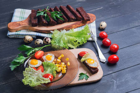 Lunch with croutons, tapas, parsley sprigs, quail eggs in shell and fried eggs, lettuce, cherry tomatoes, corn grains on dark wooden backgroundの写真素材