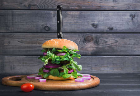 Tall sandwich hamburger deluxe with fresh green salad, tomatoes, onions on a gray wooden background, bears the knife, empty place for textの写真素材