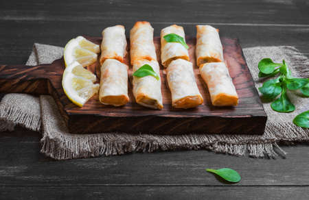 Little spring rolls on a cutting board on sacking on wooden black worn surface, lettuce, lemon slicesの写真素材