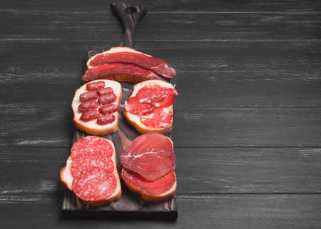 Several sandwiches with smoked and jerked meat and sausage on bread on a dark black surface, empty place for textの写真素材