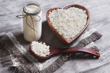 Granulated cottage cheese in a wooden bowl in the form of heart dairy cream milk jar, burlap background on a light surfaceの写真素材