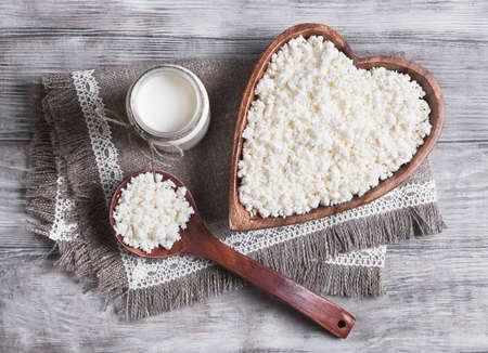 Granulated cottage cheese in a wooden bowl in the form of heart dairy cream milk jar, burlap background on a light surfaceの写真素材