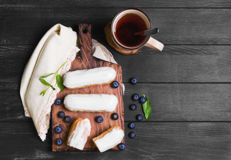 Morning breakfast Cakes French eclairs with white icing, mint, drink on a cutting board on a dark black wooden background on a linen napkin with lace, top view, empty place for text, recipeの写真素材