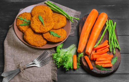 Orange carrot cutlets for vegetarian health food, peeled carrots, sliced carrots for the cutlets into strips, silver forks, asparagus, fresh herbs on meshkvoine on a dark black wooden background in rustic styleの写真素材