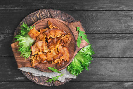 Closeup of fried bacon strips on brown wooden plate, burlap cloth, green lettuce leaves on a dark black background in rustic style, top viewの写真素材