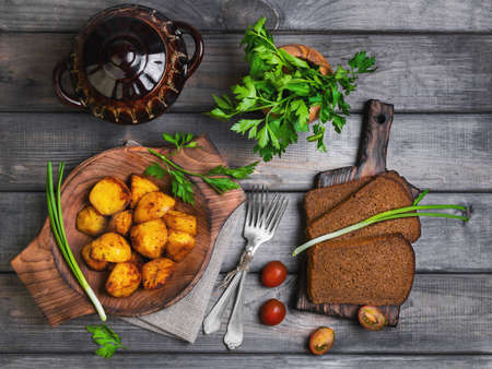 Baked potatoes roasted pieces, a ceramic pot saucepan of roasted pieces baked potatoes, rye bread, green onion, cherry tomatoes, silver fork, parsley, on a gray wooden rustic backgroundの写真素材