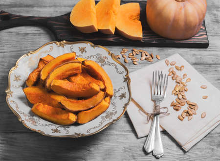 Baked pumpkin pieces on white vintage porcelain plate, raw pumpkin pieces cut for baked, napkin tissue, pumpkin seeds on a bright white background woodenの写真素材