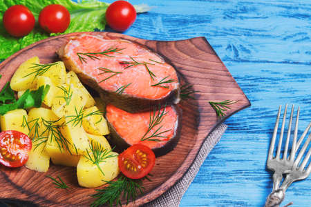 Baked fish with potatoes salmon steak fillet red fish, potato slices, parsley, dill, lettuce, cherry tomatoes on sacking on a blue wooden backgroundの写真素材