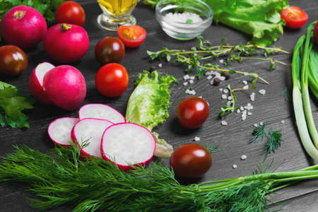 Ingredients for the preparation of fresh vegetarian vegetable salad, vegetables radishes, cherry tomatoes, lettuce, green salad dill, parsley, allspice, salt, olive oil and thyme on a dark black wooden backgroundの写真素材