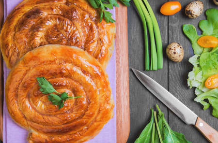 spiral filo pies burek, Greek Bougatsa, lettuce, green onions, parsley, yellow cherry tomatoes, quail eggs in a wooden bowl on a napkin on a dark black background in rustic style, top viewの写真素材