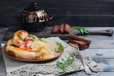 Sauerkraut with fried Munich sausages, pickled mushrooms, fresh green lettuce, silver forks, ceramic kstrblya, rye bread croutons on linen lace napkin on gray wooden background in rustic styleの写真素材
