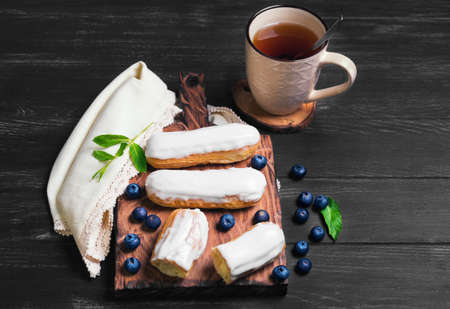 Morning breakfast Cakes French eclairs with white icing, with a special cream eclairs, mint, berries blueberries, drink on a cutting board on a dark black wooden background on a linen napkin with laceの写真素材