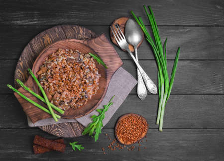 Buckwheat cereal with roasted mushrooms in a wooden plate, raw buckwheat to cereal, asparagus, pot pottery, silver fork and spoon, green lettuce leaves on a dark black background wooden surfaceの写真素材