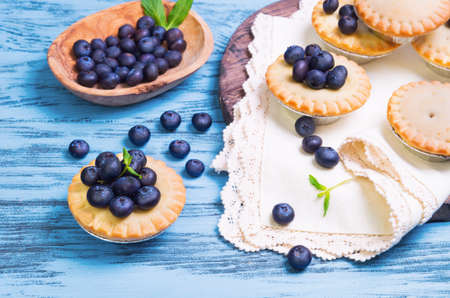 Closed berry tartlets baskets Cake with berries blueberries into molds foil for tartlets on a round wooden board, cloth with lace, mint leaves, blueberry berries in a bowl, on blue surfaceの写真素材