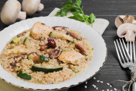 Rice cooked risotto with chicken meat and mushrooms and vegetables ingredients for risotto cherry tomatoes, zucchini squash, mushrooms, champignons on a white porcelain plate, silver fork on a dark black wooden backgroundの写真素材