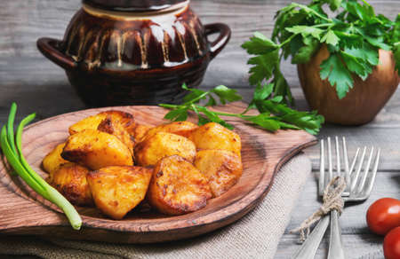 Baked potatoes roasted pieces, a ceramic pot saucepan of roasted pieces baked potatoes, rye bread, green onion, cherry tomatoes, silver fork, parsley, on a gray wooden rustic backgroundの写真素材