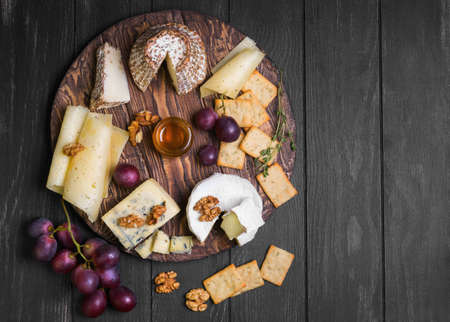 Assorted cheeses on round wooden board plate Camembert cheese, grated bark of oak, hard cheese slices, walnuts, grapes, crackers, bread, thyme, dark black wood background, top viewの写真素材