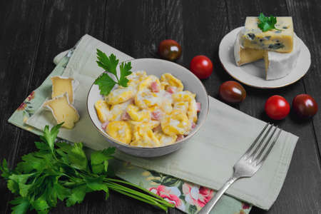 Tortellini with cheese sauce, ham, tortellini ingredients for cherry tomatoes, Camembert cheese, blue cheese, parsley, a silver fork on a dark black wooden backgroundの写真素材