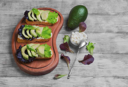 Three sandwiches with avocado, cottage cheese ricotta cheese, lettuce, ingredients for sandwiches cream cheese in bowl, creeping,  fruit avocado on a cutting board on a light white wooden backgroundの写真素材