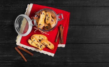 Italian biscotti cookies in a glass jar, cinnamon sticks, cookies biscotti scattered on a red lacy napkin on a dark black wooden background, top viewの写真素材