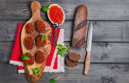 Small canape sandwiches with fish red caviar, leaves green salad, rye bagget, fish red caviar in white plate, red linen napkin lace on cutting board olive wood on gray background wooden surfaces, top viewの写真素材