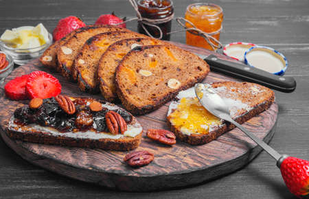 Sweet dessert sandwiches crostini with lemon jam, plums jam, nuts pecans, hazelnuts, almonds, strawberries, rye bread with prunes and dried apricots for sandwiches, butter, cutting board on black wooden backgroundの写真素材