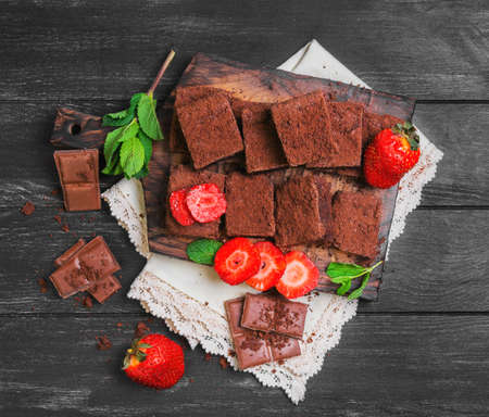 Pieces of chopped chocolate cake brownie with nuts, chocolate bars, leaves and sprigs of mint, crumb cake brownies, strawberry berries on a cutting board on a black wooden backgroundの写真素材