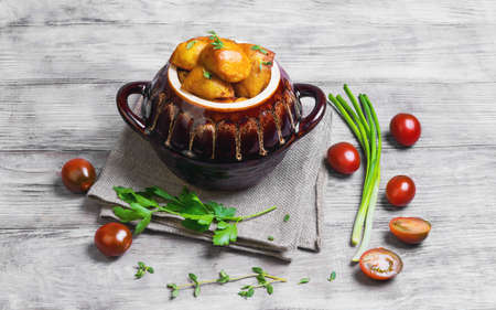 Baked potatoes in the oven in a special ceramic pot for roasting potatoes, chives, thyme, parsley, burlap cloth on a bright white background woodenの写真素材