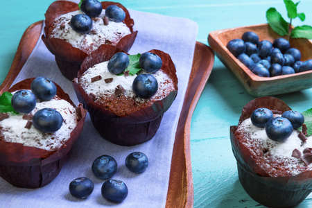 Muffins berries blueberry cupcakes in paper forms for muffins, on top of a bowl of blueberries berries, sprigs of mint and grated chocolate shavings on a wooden tray on a green backgroundの写真素材