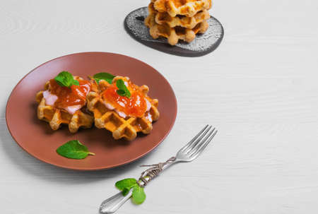 Freshly soft thick Belgian waffles with apricot jam, mint leaves, cream, silver fork on a white plate on a brown wooden backgroundの写真素材