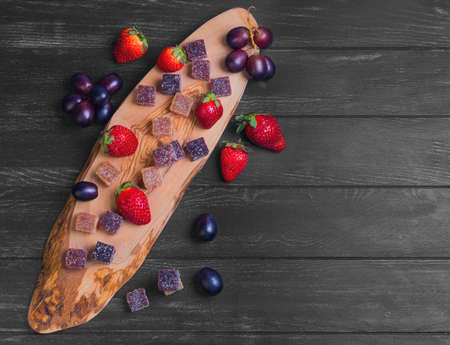 Fruit and berry jellies marmalade, purple grapes, red strawberries on board wood of olive, three cubes fruit jelly marmalade on black wooden background, top viewの写真素材