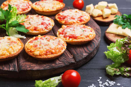 Small pizza with mozzarella cheese, sausage, ham, parsley, lettuce, cherry tomatoes in olive wood planks on dark black wooden backgroundの写真素材