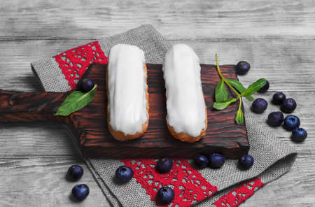 Two French Cake eclairs with white icing on wooden board, burlap cloth with red lace, mint leaves, Blueberry berries for filling eclairsの写真素材