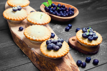 Closed berry tartlets baskets Cake with berries blueberries into molds foil for tartlets on olive wooden board, mint leaves, blueberry berries in bowl, on black surfaceの写真素材