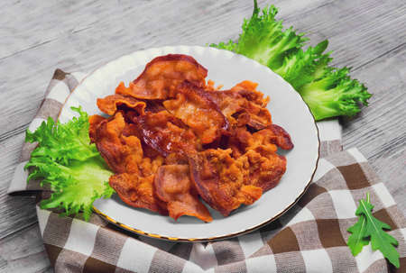 Closeup of fried bacon strips on white plate, cloth, green lettuce leaves on light whit background in rustic styleの写真素材