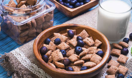 Multigrain healthy cereal corn pads chocolate in white bowl for milk breakfast, glass of milk, cereal bank, cereal is scattered on table, blueberries, bilberries, blue wooden backgroundの写真素材