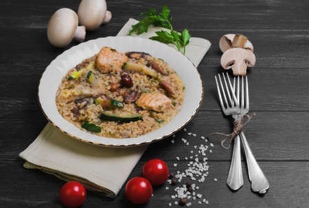 Rice cooked risotto with chicken meat and mushrooms and vegetables ingredients for risotto cherry tomatoes, zucchini squash, mushrooms, champignons on white porcelain plate, silver fork, dark black wooden backgroundの写真素材