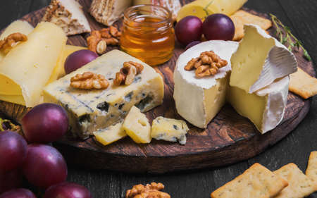 Assorted cheeses on round wooden board plate. Camembert cheese, blue cheese, goat cheese in the grated bark of oak, hard cheese slices, walnuts, grapes, crackers, bread, thyme, honey sauce, dark black wood backgroundの写真素材