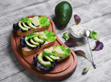 Three sandwiches with avocado, cottage cheese ricotta, lettuce, ingredients for sandwiches cream cheese in bowl, creeping,  fruit avocado on cutting board on light white wooden backgroundの写真素材
