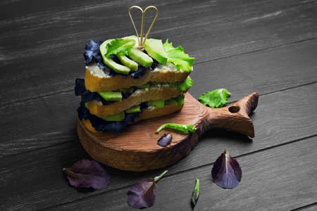 High large layered sandwich with avocado, lettuce, cheese ricotta on acutting board on dark black wooden backgroundの写真素材